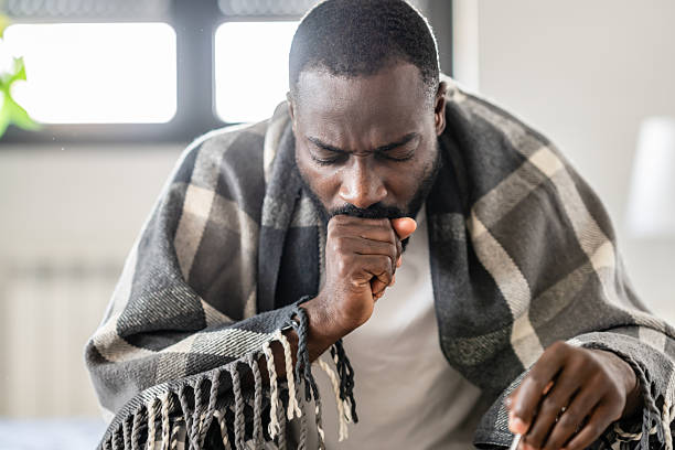 istock Sick man wrapped in a plaid blanket, coughing into his fist while sitting indoors, looking unwell and fatigued. 2204568789