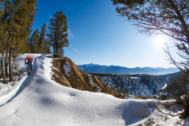 A woman rides down a hill along the edge of a river canyon during a winter fat bike ride on a sunny morning near Invermere, British Columbia, Canada. She wears a cycling helmet, warm winter clothing, and a backpack. Fat bikes are mountain bikes with large wheels and tires for riding on the snow.