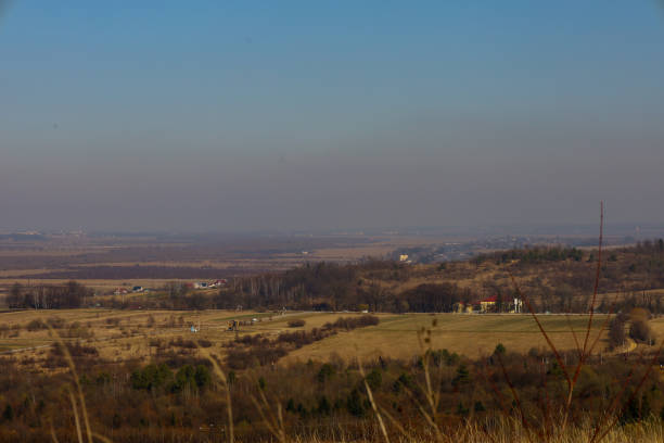 istock Panoramic view of a rural area from a hill. In the foreground, dry grass and tree branches create a frame for the shot. In the distance, fields, forest belts, and small villages are spread out. 2204571389