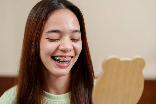 istock Asian young woman smiling joyfully while looking at a bear-shaped wooden mirror during a relaxing holiday. Natural look, long brown hair, wearing green long-sleeve shirt and dental braces. 2204573334