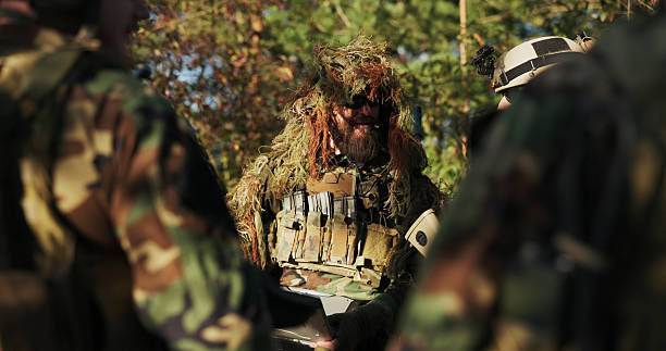 istock Soldiers meet at the base to plan tactics, while the commander in grass camouflage analyzes topography on a computer, discussing strategic movements with the team. 2204573647