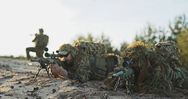istock A tactical soldier, lying in concealment, opens the flap of his binoculars, monitors enemy forces, and positions his gun on a stand, fully prepared for a territorial defense training exercise. 2204577094