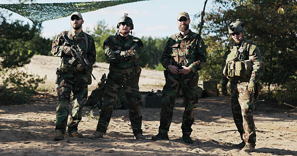 istock Four army men in camouflage uniforms stand ready, inspecting weapons, unlocking short pistols, adjusting rifles, and ensuring all gear is secured in holsters and belts. 2204578631