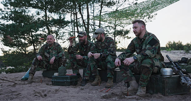 istock Soldiers in camouflage uniforms unwind at the base after their shift, sitting outdoors on crates, building a campfire, and skewering sausages on sticks for a meal in the field. 2204580855