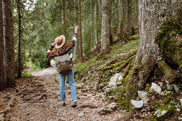 A young tourist with a hiking backpack walks along a hiking path in a mountain forest. Happy woman enjoys the weather outdoors. Hiking, travel concept. Active lifestyle.