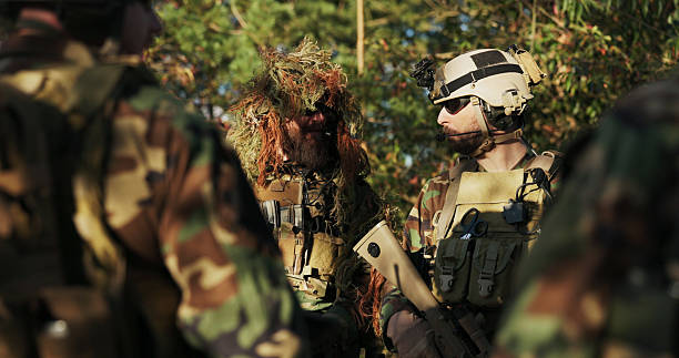 istock Troops assemble at the base to plan their tactical approach, as a soldier in grass camouflage holds a computer and discusses strategy with the commander. 2204582731