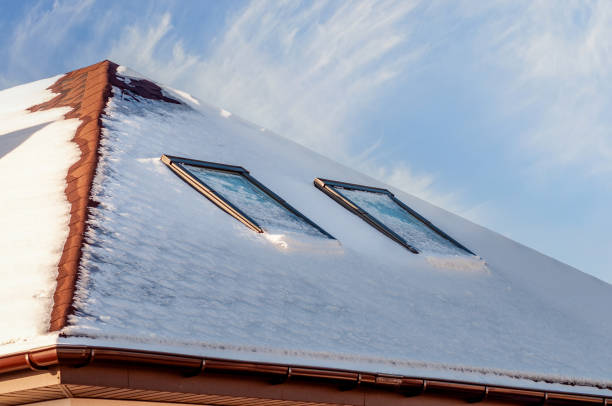 A rooftop covered in snow with two skylights peeking through the wintry blanket against a backdrop of a partly cloudy blue sky.