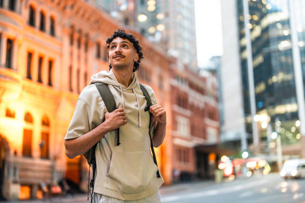A Hispanic male in casual attire and a backpack explores Sydney streets at dusk, surrounded by bright urban lights and historic architecture.