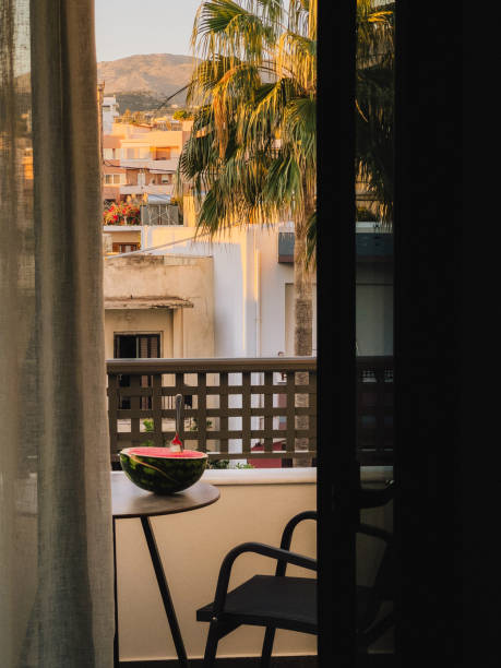 istock Half watermelon with spoon on small table on balcony overlooking city with palm trees and mountains in background during summer sunset, suggesting relaxing holiday atmosphere. leisurely vacation mood 2204593336