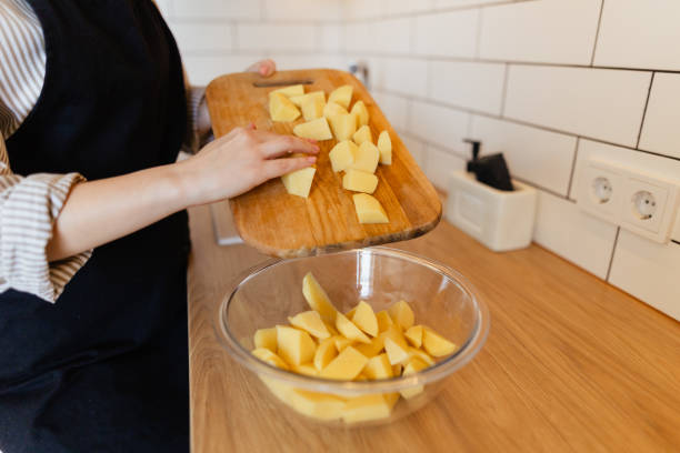 Close-up of a hand in a black glove using a sharp knife to slice a peeled potato into even pieces on a wooden board, showcasing meal preparation