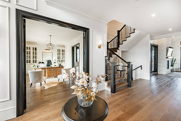 Beautiful large and luxurious estate home interior front door entry hallway foyer with large glass and black wood door expansive entry with a round table containing an arrangement of dried flowers in a bouquet. The view from the front is to a home office and a casual sitting room with blue velvet sofas. large wooden barn doors lead to another room there is a staircase and steps going up.