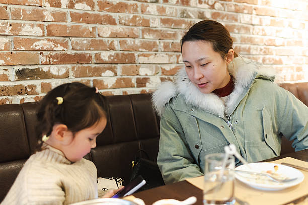 A Chinese mother and daughter are talking in a restaurant while waiting for their dinner. A little girl plays with the phone while her mother talks to her