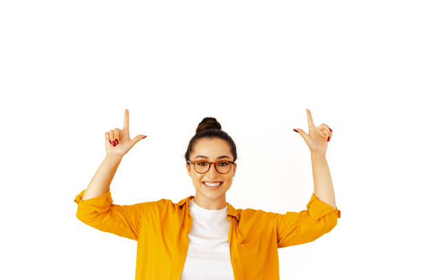 Happy caucasian or arabian woman in a yellow shirt with glasses, points upward with both hands to an empty copy-space for presentation or advertising, stand on isolated white background, smiling stock photo