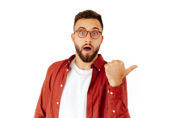 Attractive excited caucasian or arabian young man in a red shirt with glasses, looks inspired and surprised while standing on isolated white background and shows finger to the side to a copy space stock photo