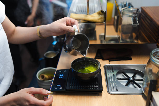 Barista carefully pouring hot water into matcha green tea powder in a bowl placed on a digital scale, preparing a matcha latte in a specialty coffee shop