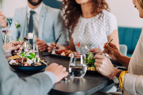 Cropped shot of group of business persons talking in a restaurant. Cheerful business people eating at the restaurant. Business team having lunch or dinner together enjoying each other company. Business discussion and fun at the lunch break. A small group of entrepreneurs having fun during a business lunch in a restaurant. Group of happy business persons communicating while enjoying in business lunch or dinner in a restaurant.