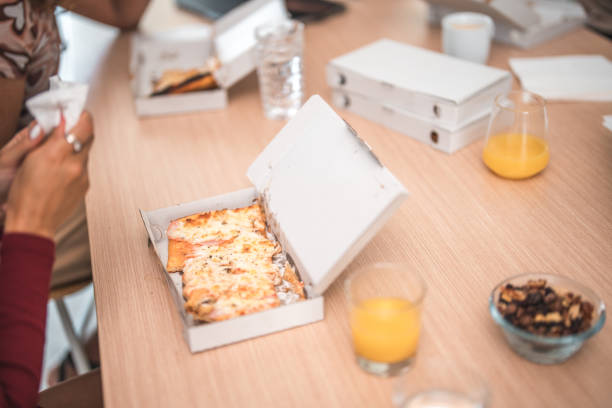 Pizza slice in a box on a table with some orange juice next to it during a lunch break at gen Zs office.