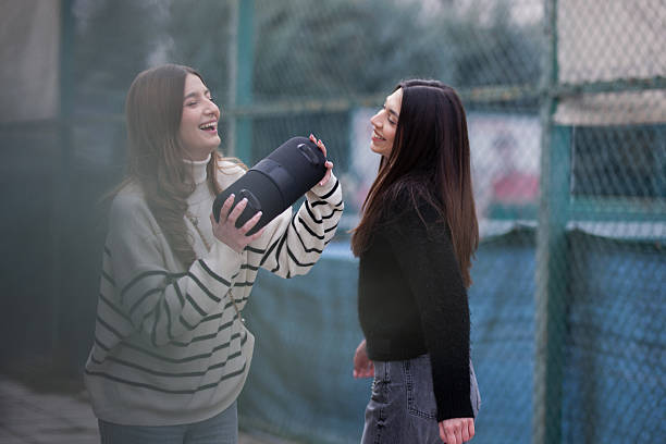 Two young women expressing joy and connection, standing outdoors on a casual day while using a portable speaker, symbolizing friendship and happiness.