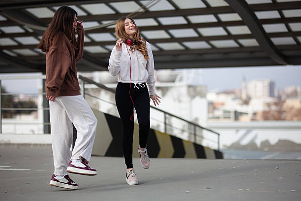 Two young women dressed casually enjoying music and conversing on a modern terrace with a city skyline in the background, expressing joy and vibrancy.