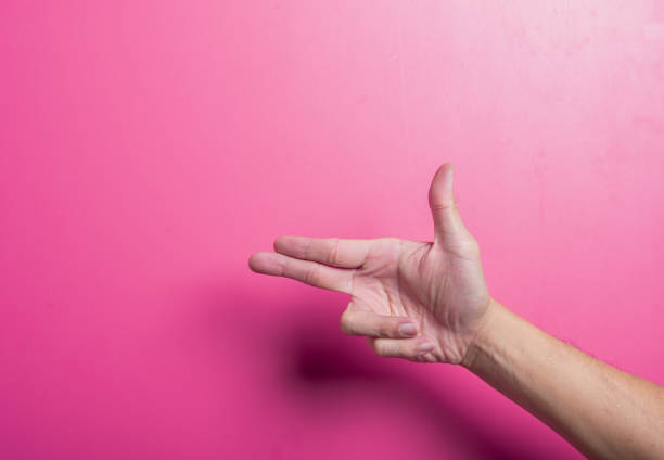 Asian hand mimicking a finger gun gesture with thumb raised and two fingers pointed forward on a vibrant pink background. Studio-lit setup ideal for playful, bold, or symbolic communication themes.