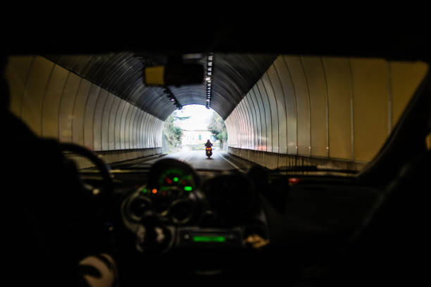 A motorcycle rides out of a dimly lit tunnel into bright sunlight. The view is from inside a vehicle, showcasing the contrast between darkness and light around the rider.