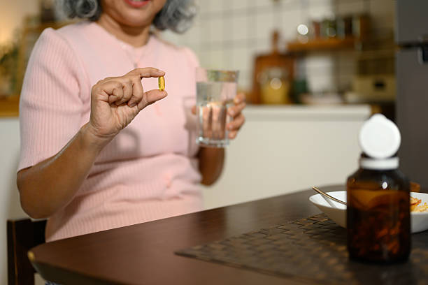 Smiling senior woman holding a fish oil capsule and a glass of water sitting in a cozy kitchen. stock photo