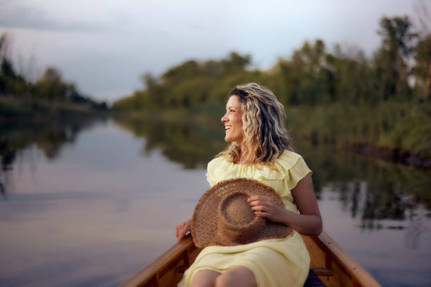 woman sits gracefully on a blue boat, feet in the water. She wears a patterned dress and appears relaxed amidst gently rippling waves and a lush green shoreline under a cloudy sky.