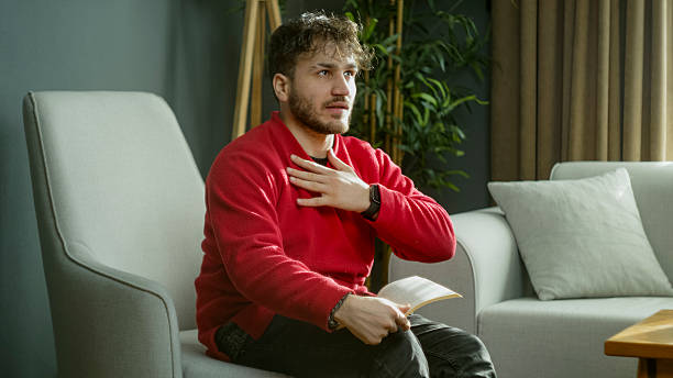 istock Young man in a red sweater laughing while reading a book, covering his face with his hand in a cozy modern living room 2204665092