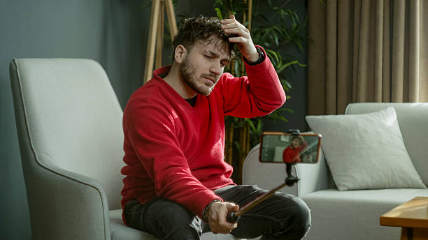istock Young man in a red sweater using a selfie stick to record a video or take a photo while sitting in a modern living room 2204666027