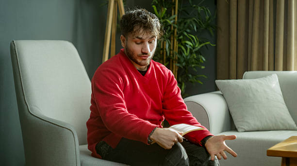 istock Man in a red sweater laughing while reading a book, covering his face with his hand in a cozy modern living room 2204666113