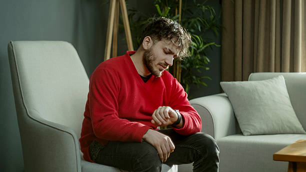 Young man in a red sweater checking the time on his smartwatch while sitting on a couch in a modern living room