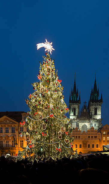 Festive atmosphere fills the air at the Prague Christmas Market, showcasing a tall, luminous tree adorned with ornaments, set against the backdrop of charming gothic architecture in the evening sky.