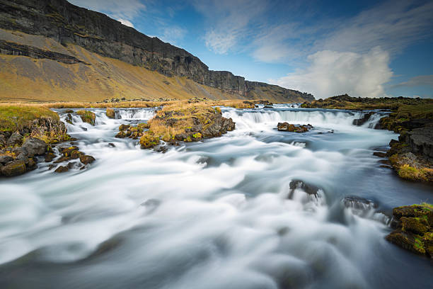 A serene waterfall flows over moss-covered rocks, surrounded by golden grass and dramatic cliffs. The clear blue sky adds tranquility to the breathtaking Icelandic scenery.