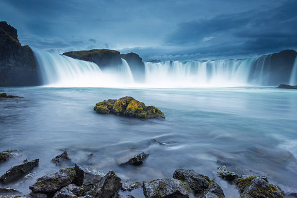 Water flows powerfully over Godafoss waterfall, surrounded by rugged rocks and a serene body of water. The dramatic clouds create a moody atmosphere in this Icelandic landscape.