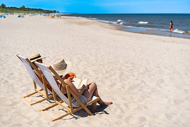istock Two mature women reading books sitting on deckchairs on beach 2204673555