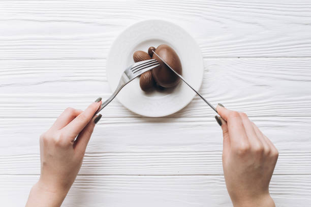 istock Female hands with knife and fork eating Easter chocolate eggs on white wooden background. 2204675431