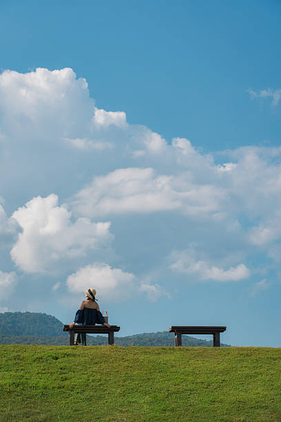 Female tourist sitting at wooden bench on lush green hill and large fluffy clouds in blue sky on sunny day