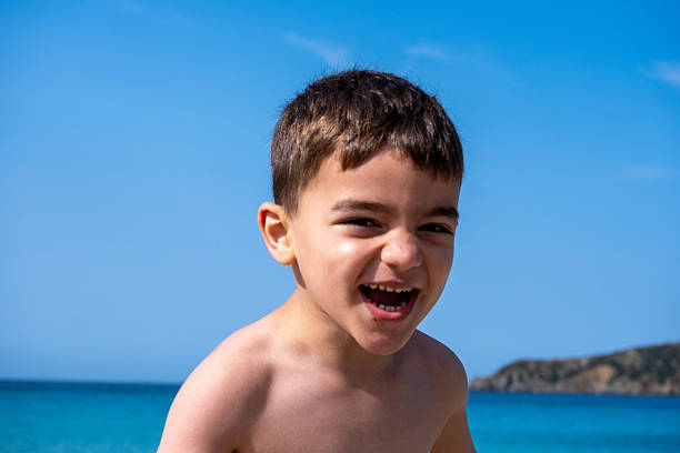 A young boy is smiling and laughing while standing on the beach. The sky is blue and the ocean is in the background