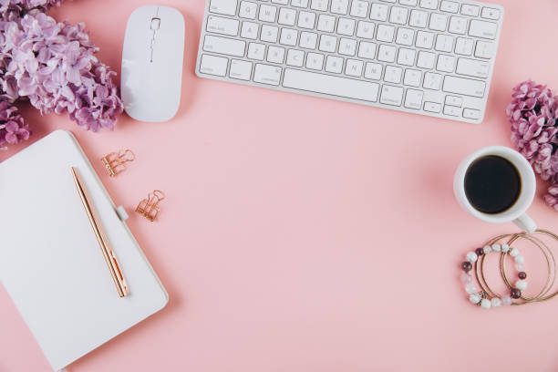 istock Spring flat lay top view home office workspace - modern keyboard, cup of coffee and notebook with lilac flowers on a pink desk background 2204681072