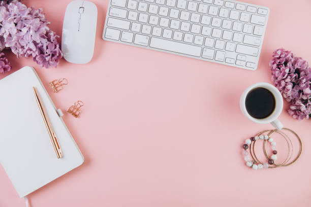 istock Spring flat lay top view home office workspace - modern keyboard, cup of coffee and notebook with lilac flowers on a pink desk background 2204681091