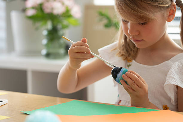 istock Cute girl with bunny ears headband painting Easter egg with paints. Preparing for Easter. 2204685967