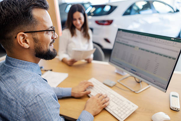 Smiling car dealer sitting at car salon and typing documents on computer while buyer reading contract