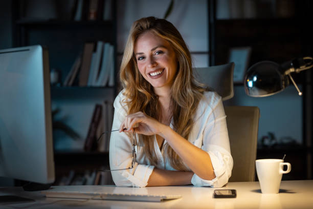 Smiling business woman working with laptop while looking at camera in modern startup office at night