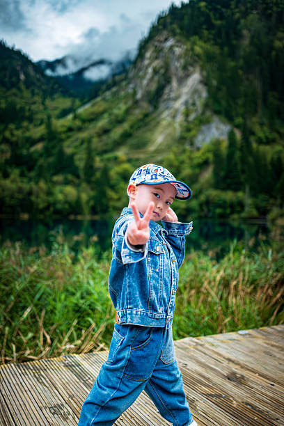 A boy stand on a wooden walkway by the lake in Jiuzhaigou. Distant green mountains reflect in the crystal-clear water, with mist swirling around, creating a poetic and picturesque scene