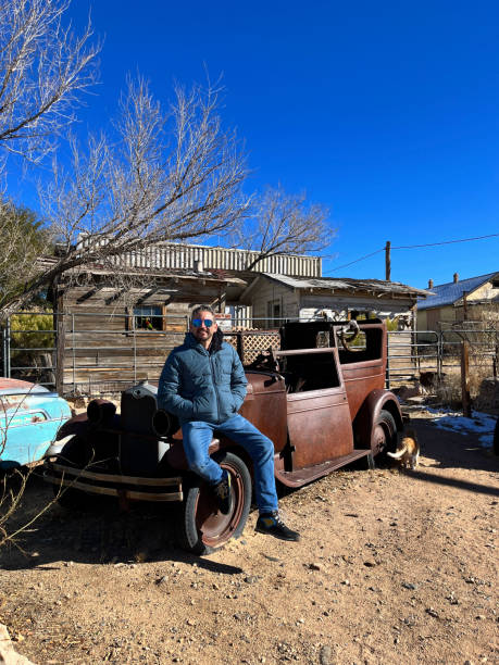 Tourist is taking a break from his road trip, posing for a photo on a rusty abandoned vintage car in the ghost town of Hackberry, Arizona, along the iconic Route 66