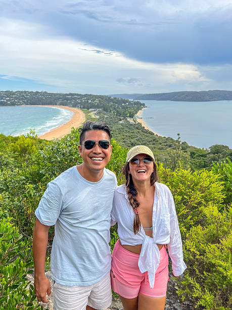 Smiling couple hiking on a rocky trail with a stunning coastal background. Happy tourists exploring nature. Friends on adventure enjoying panoramic ocean and landscape views.