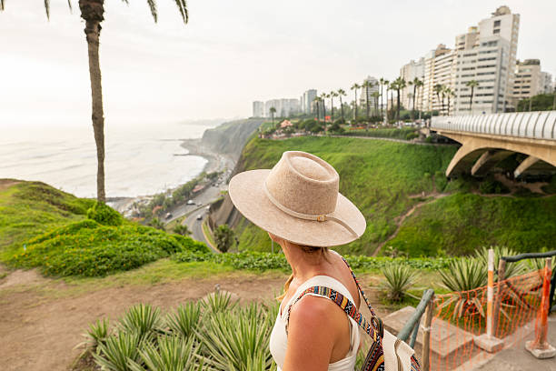 Woman in hat gazing at ocean from cliff, lush green landscape and city skyline. Traveler exploring scenic viewpoints. Perfect for tourism and adventure marketing.
