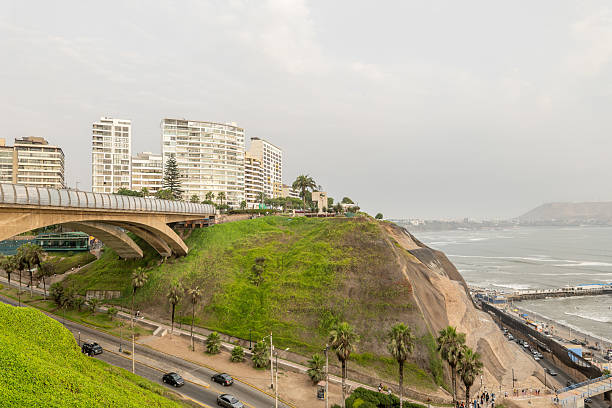 Modern bridge curving over lush green hillside in coastal cityscape. Tall buildings overlook the ocean. Scenic urban environment with ocean waves hitting the shore.
