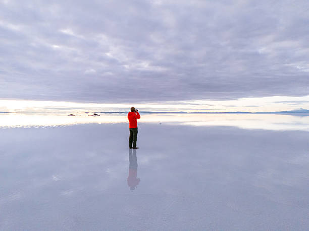 A lone photographer in a red jacket takes photos on expansive salt flats. A serene scene of nature's beauty, clouds reflecting on the vast, mirrored surface.