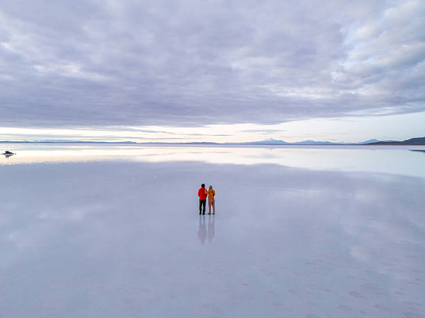 Couple embraces on the expansive salt flats, reflecting a serene overcast sky. Two people admiring nature’s beauty.